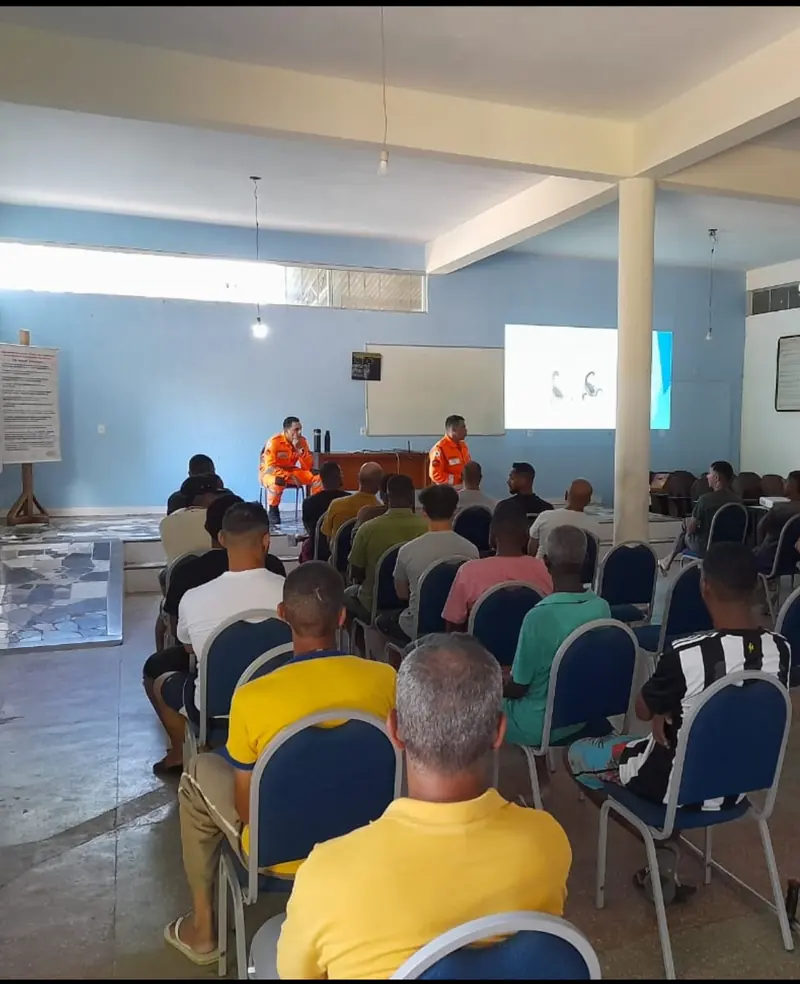 Salão da Colônia com acolhidos sentados, vistos de costas, assistindo a uma palestra de dois bombeiros uniformizados de laranja, com projeção na parede.