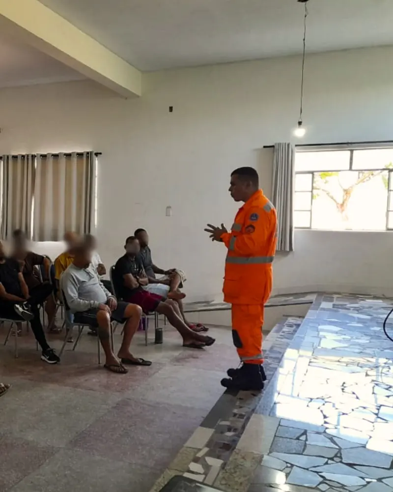 Bombeiro uniformizado de laranja em pé, falando a um grupo de acolhidos sentados num salão; os rostos dos acolhidos estão desfocados.