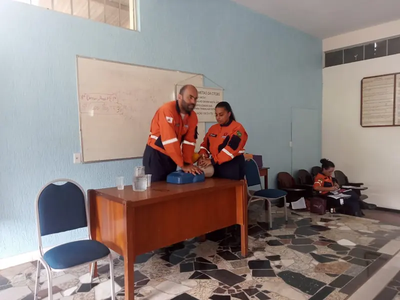 Dois bombeiros uniformizados demonstram a massagem cardíaca em um boneco sobre uma mesa, diante de um quadro branco.