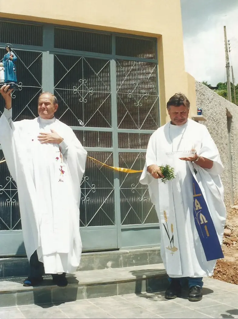 Dois religiosos de vestes brancas diante de um portão de ferro com fita amarela, um deles erguendo uma imagem de santa.