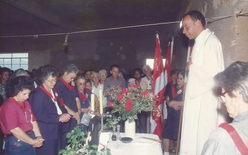 Celebração religiosa dentro de uma construção ainda sem acabamento, com fiéis em oração, flores, velas e bandeiras vermelhas e brancas.