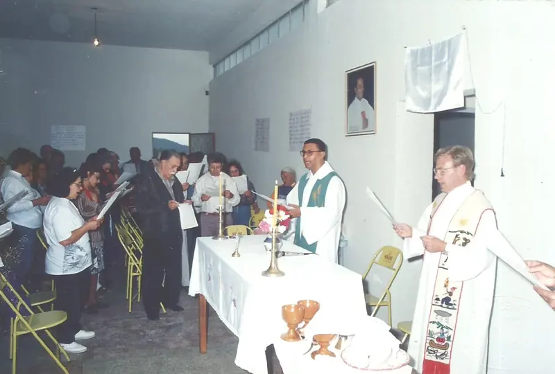 Celebração religiosa em uma sala com fiéis em pé segurando folhas, dois religiosos de vestes brancas junto à mesa do altar.