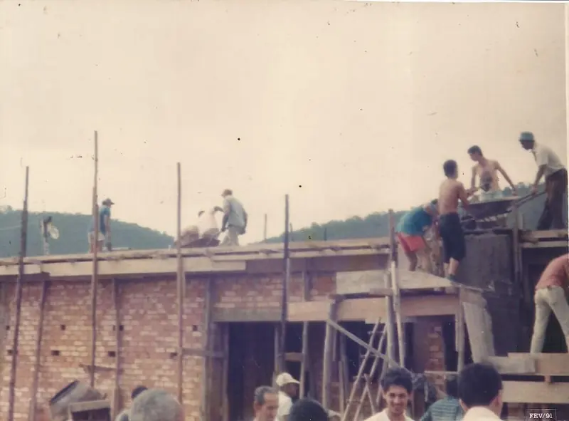 Obra de tijolos aparentes com operários no telhado e em andaimes, sob céu claro, com carimbo FEV/91 no canto.