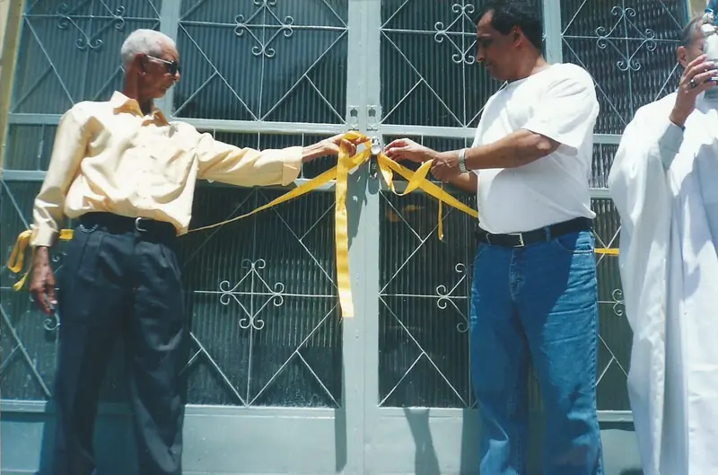 Dois homens seguram uma fita amarela diante de um portão de ferro, ao lado de um religioso de veste branca.