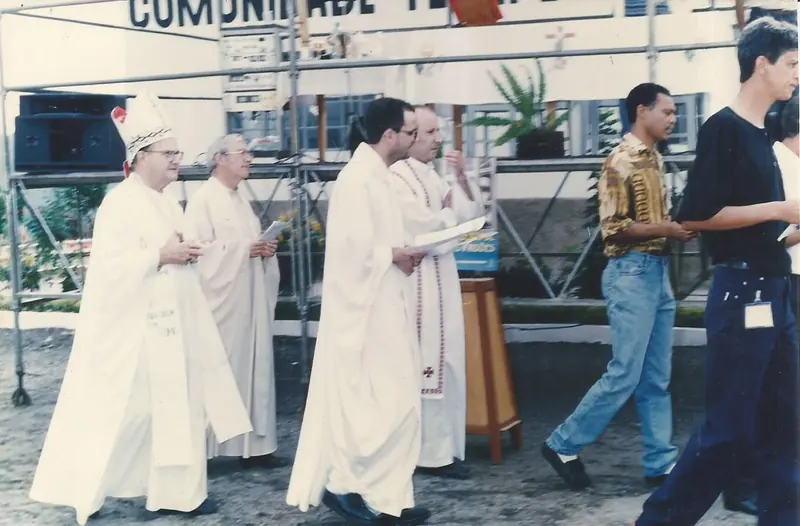 Religiosos de vestes brancas caminhando em procissão diante de uma estrutura de andaimes, com a faixa “Comunidade Te…” ao fundo.