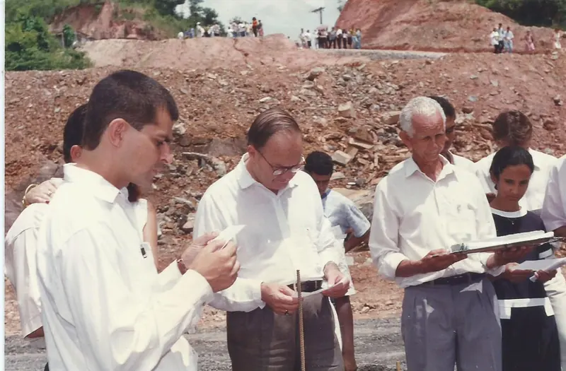 Homens de camisa branca lendo papéis em um terreno de terra vermelha, com pessoas ao fundo sobre o barranco.
