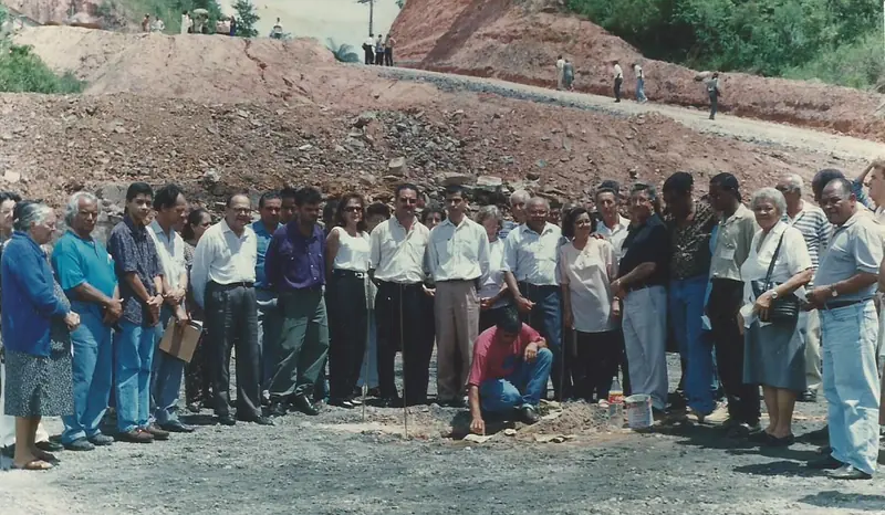 Grupo grande de pessoas em pé em um terreno de terra e brita, ao redor de um homem agachado no chão.