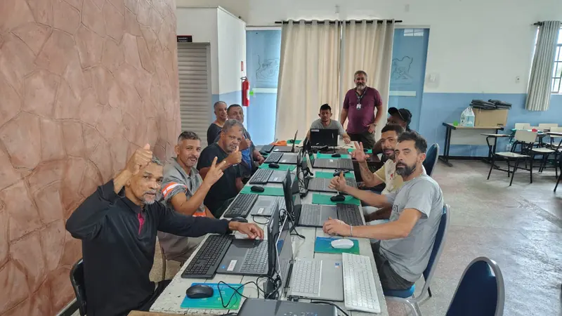 Turma do curso de informática numa sala, sentada ao longo de uma mesa comprida com notebooks; os alunos sorriem e fazem sinal de positivo para a câmera, e um homem aparece em pé ao fundo.