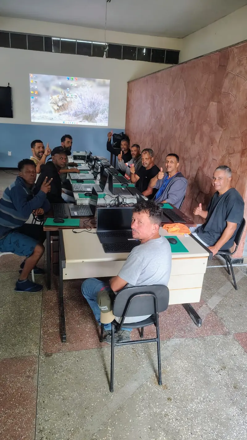 Alunos do curso de informática sentados ao redor de mesas com notebooks, diante de uma tela de projetor que mostra a área de trabalho do computador.