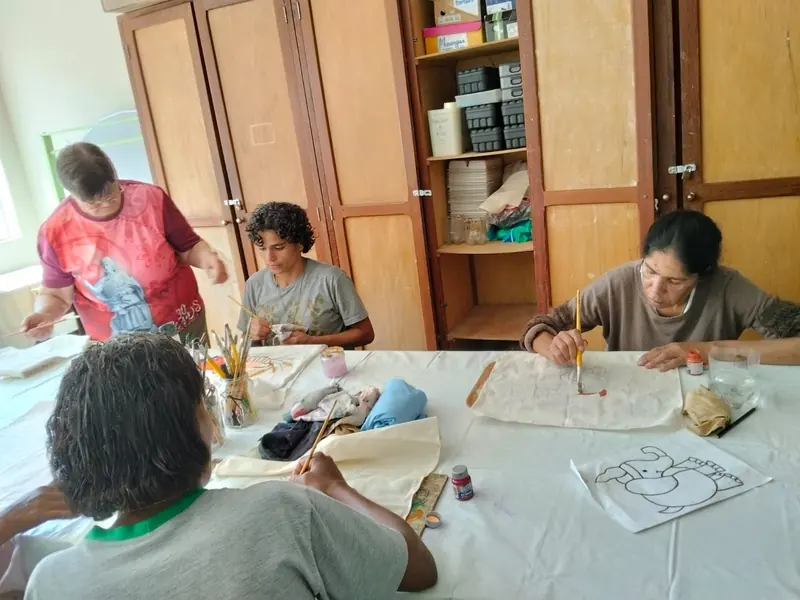 Quatro mulheres ao redor de uma mesa coberta com toalha branca pintando desenhos em panos de tecido, com pincéis e tintas, em uma sala com armários de madeira.