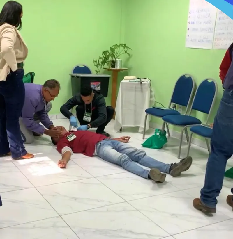 Participantes do curso de primeiros socorros praticando o atendimento a uma pessoa deitada no chão de uma sala de paredes verdes.