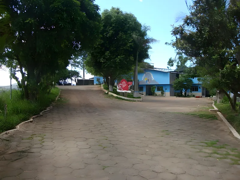 Foto real: alameda calçada de entrada da Colônia, com árvores dos dois lados e, ao fundo, o prédio azul e o coração vermelho de boas-vindas