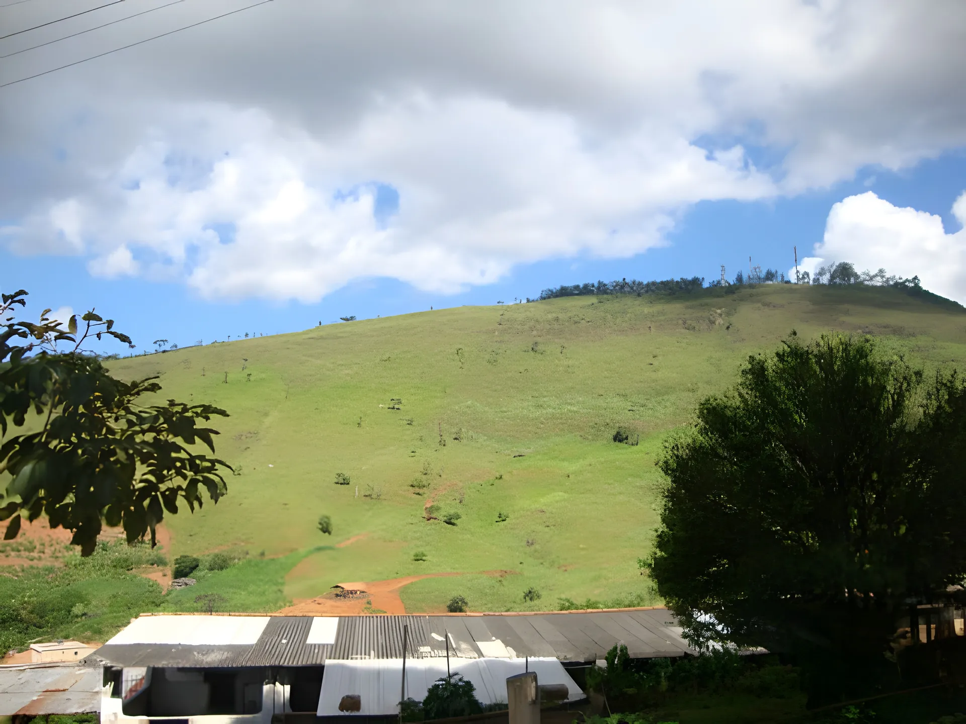 Colina verde sob céu azul com nuvens, vista da Colônia, com telhados no pé do morro