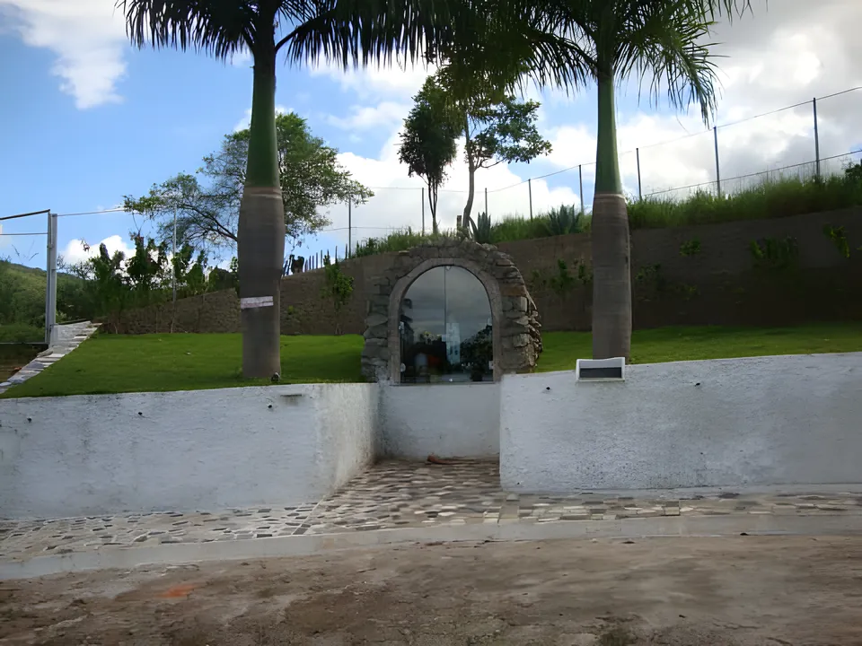 Gruta com imagem religiosa em um muro branco, entre palmeiras, na Colônia